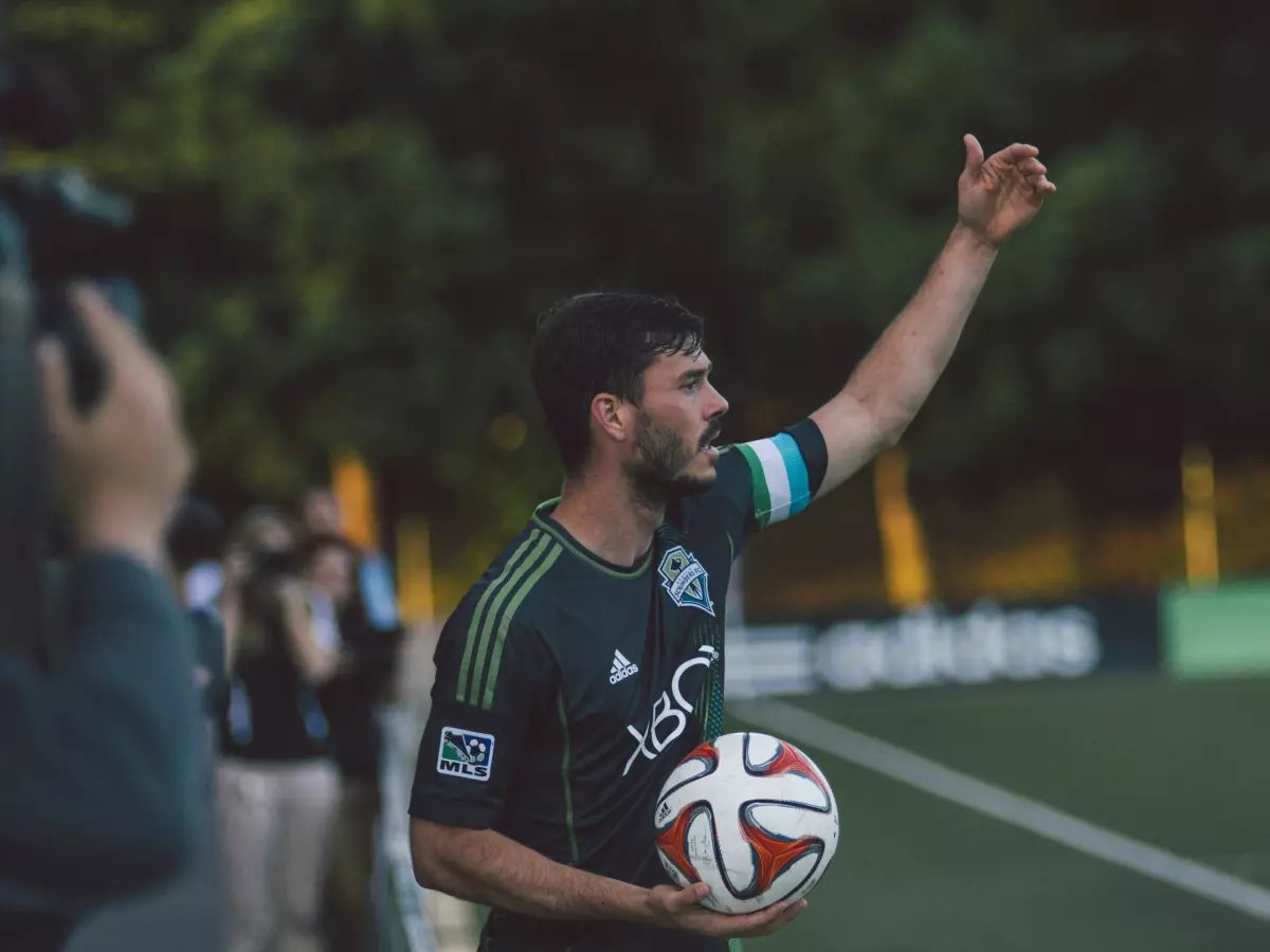 A soccer player holding a soccer ball, with a blurred background of other people and a green field.