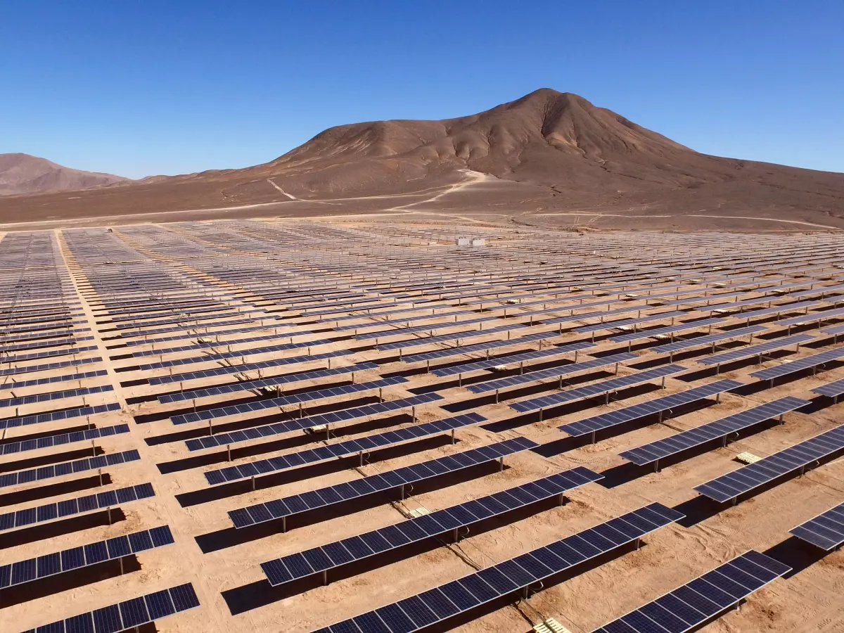 An expansive desert solar farm stretches across a vast landscape under a clear blue sky, with mountains in the distance.