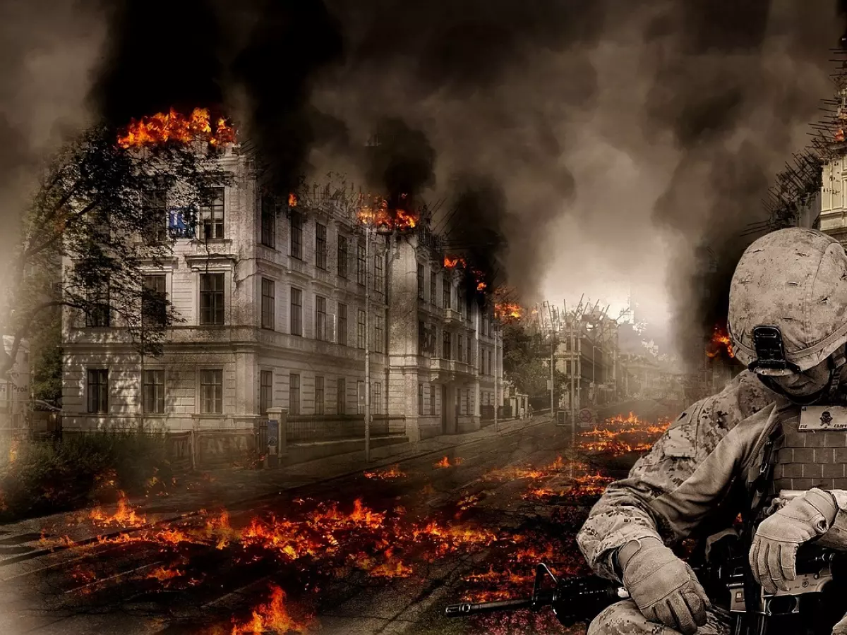 A soldier in a military uniform is sitting on a street in a war-torn city. The city is engulfed in flames, and the soldier is looking down at his rifle.