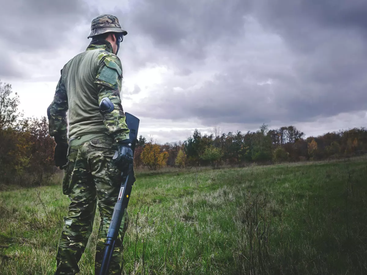 A lone soldier stands in a field with a rifle, looking out over a forest. The sky is cloudy and ominous, suggesting a sense of danger or uncertainty.