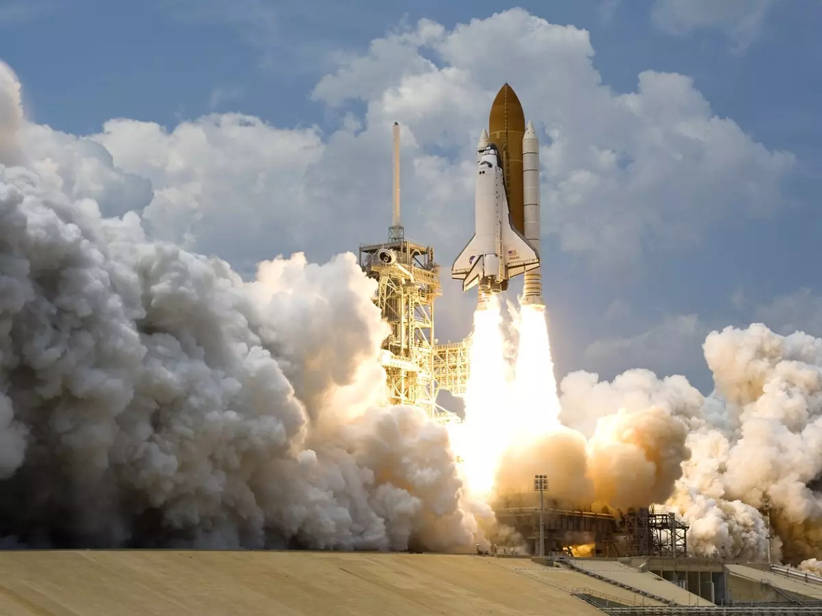 A space shuttle launches from a platform, with a bright flame and smoke behind it.