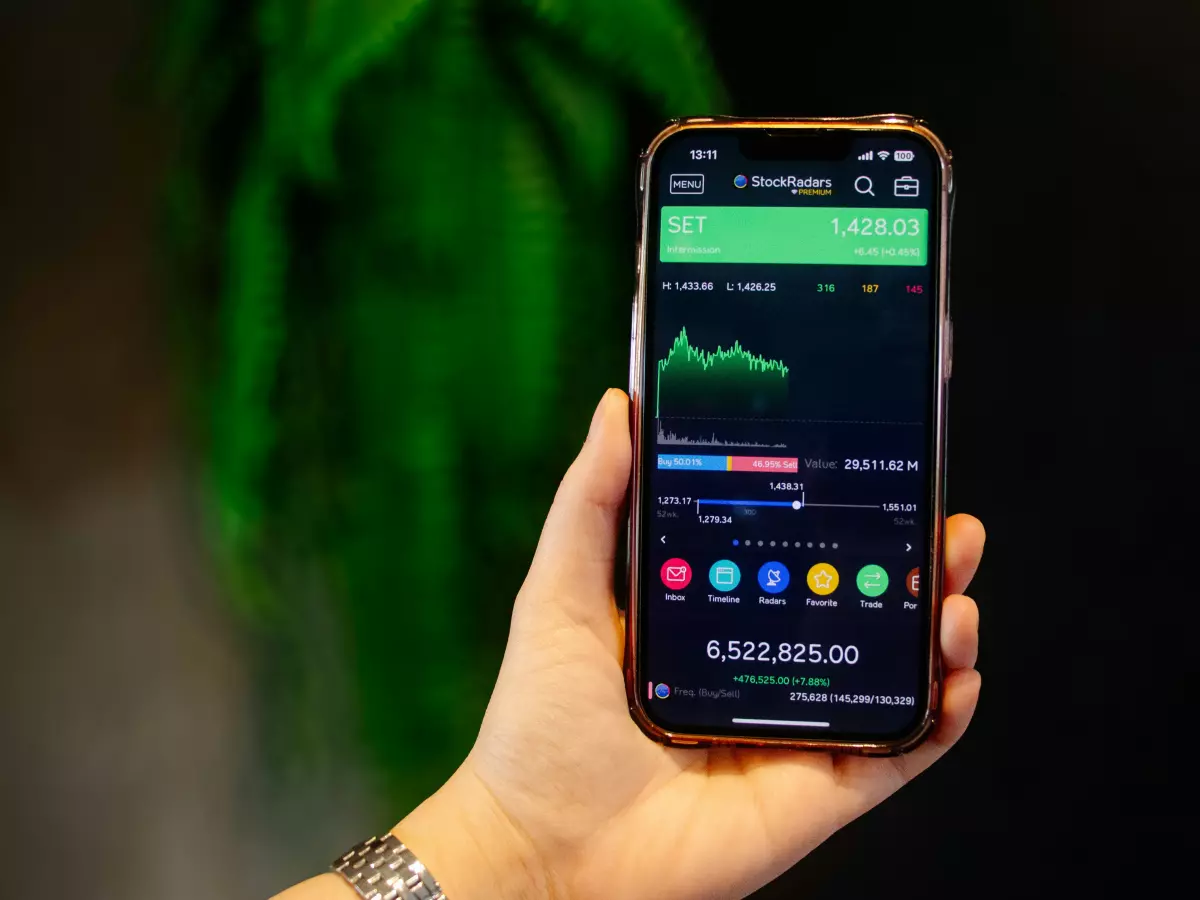 A hand holding a smartphone displaying a stock market graph. The phone is in the foreground, with a blurred green plant in the background.