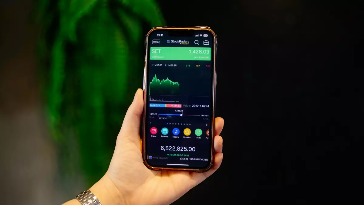 A hand holding a smartphone displaying a stock market graph. The phone is in the foreground, with a blurred green plant in the background.