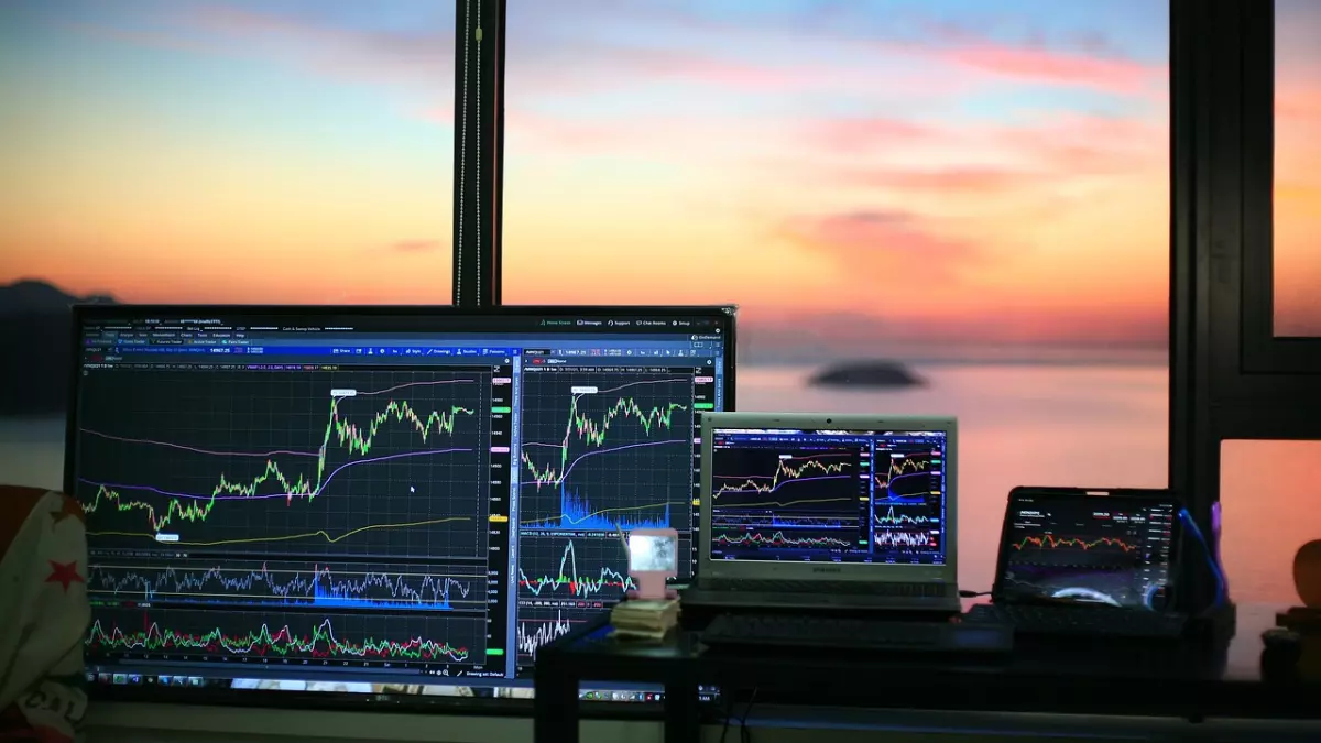 Three computer screens and a laptop are set up on a table, all showing stock market charts. The screens are facing a window with a view of a sunset over the ocean.