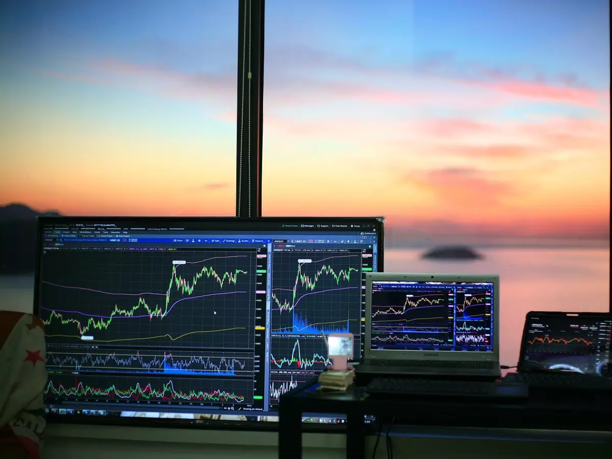 Three computer screens and a laptop are set up on a table, all showing stock market charts. The screens are facing a window with a view of a sunset over the ocean.