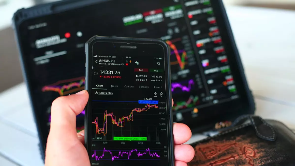 A close-up shot of a hand holding a smartphone displaying stock market data, with a laptop screen in the background showing similar data.