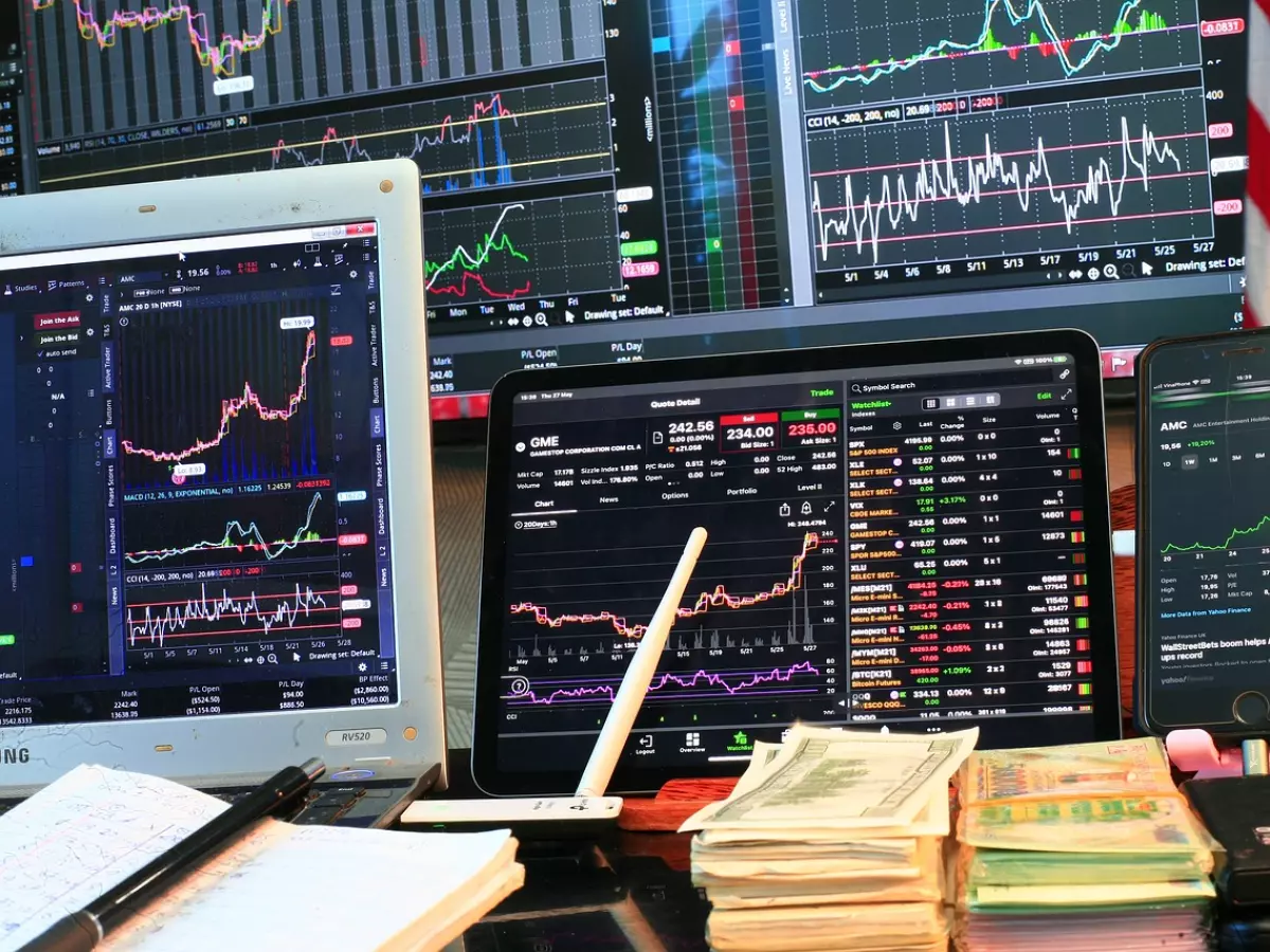 Close-up view of multiple computer screens displaying financial data, including stock charts and financial indicators. There are also some paper bills and a smartphone in the foreground.