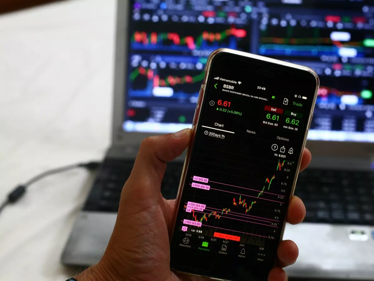 A hand holds a smartphone showing a crypto trading app, with a laptop in the background.