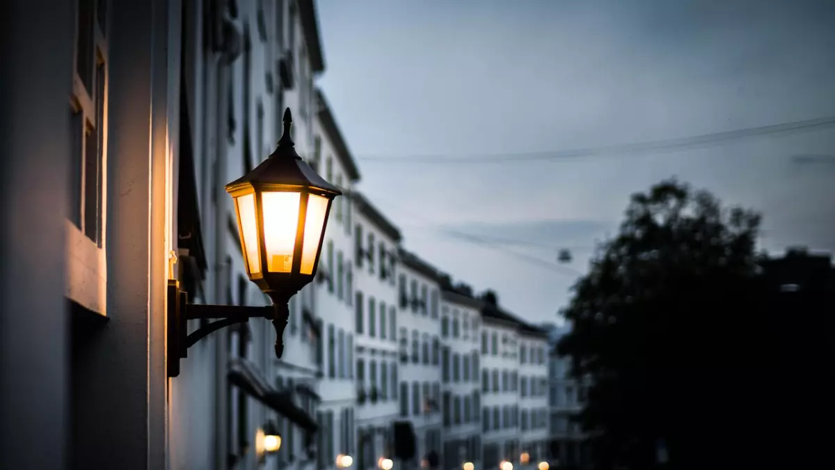 A street lamp with a warm glow on a street at dusk, highlighting the lamp and blurring the street and buildings.