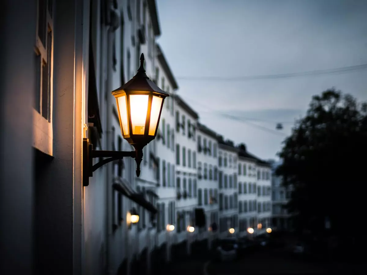 A street lamp with a warm glow on a street at dusk, highlighting the lamp and blurring the street and buildings.