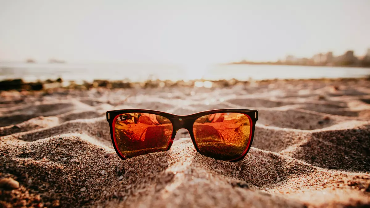 A pair of sunglasses lie on a sandy beach, with a blurred background of a coastline and the setting sun. 