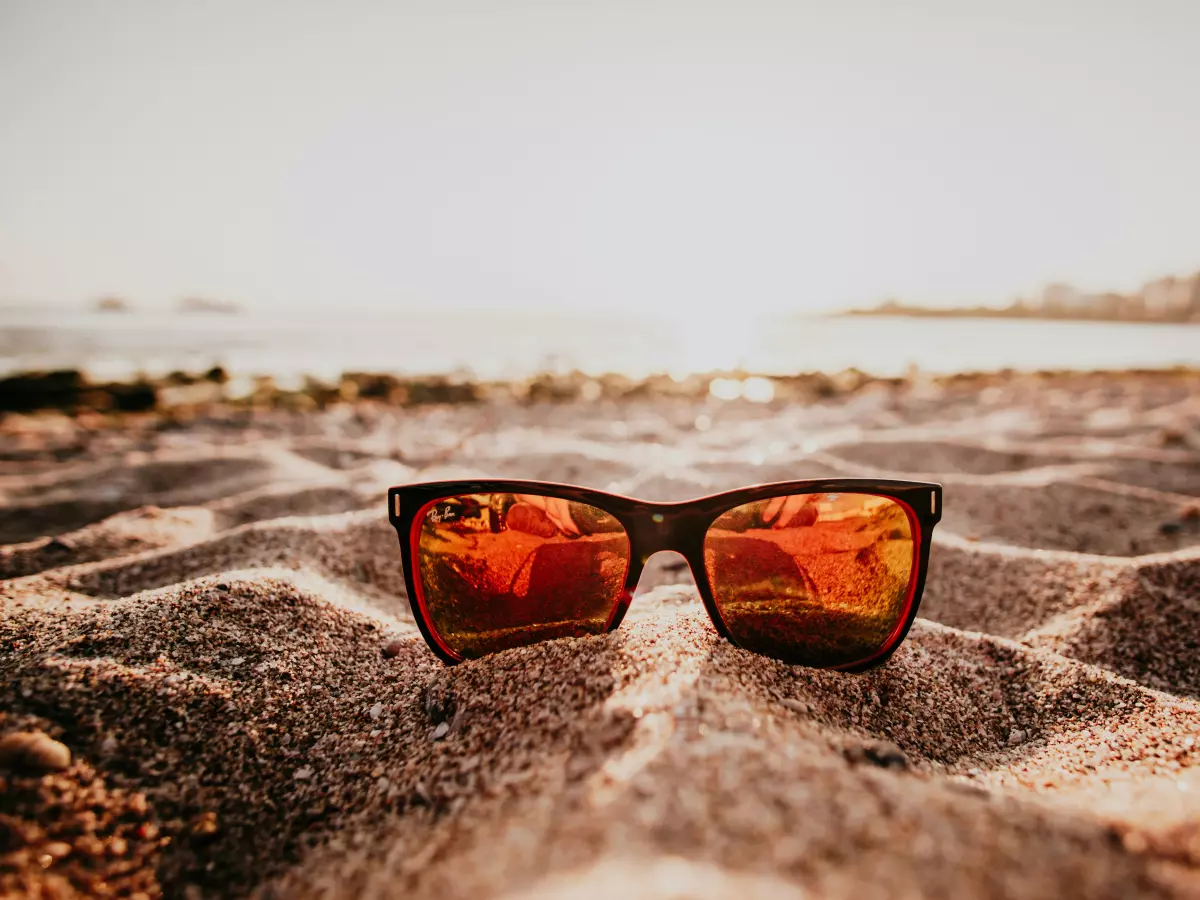 A pair of sunglasses lie on a sandy beach, with a blurred background of a coastline and the setting sun. 