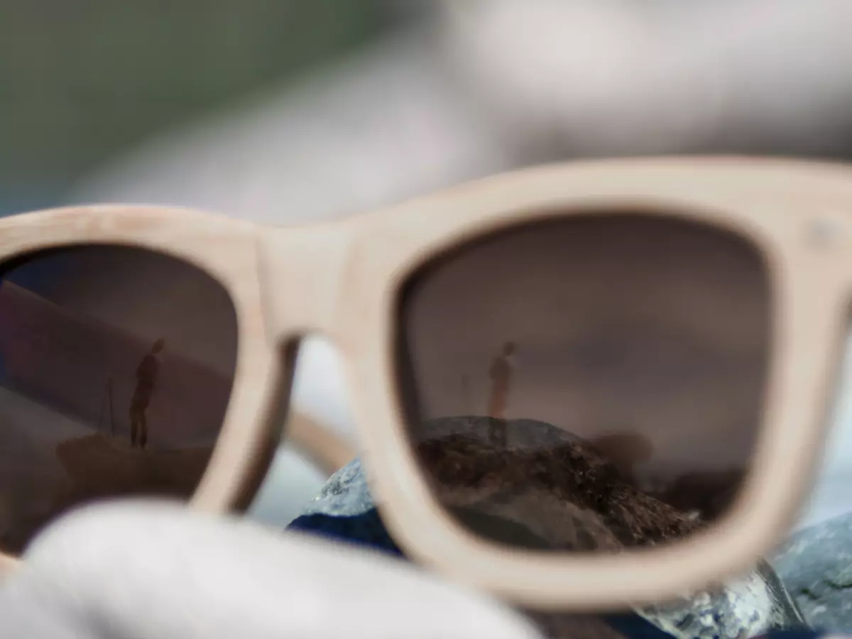 Close-up shot of a pair of wooden sunglasses, with a reflection of a person standing in the background. The sunglasses are resting on a surface covered in pebbles.