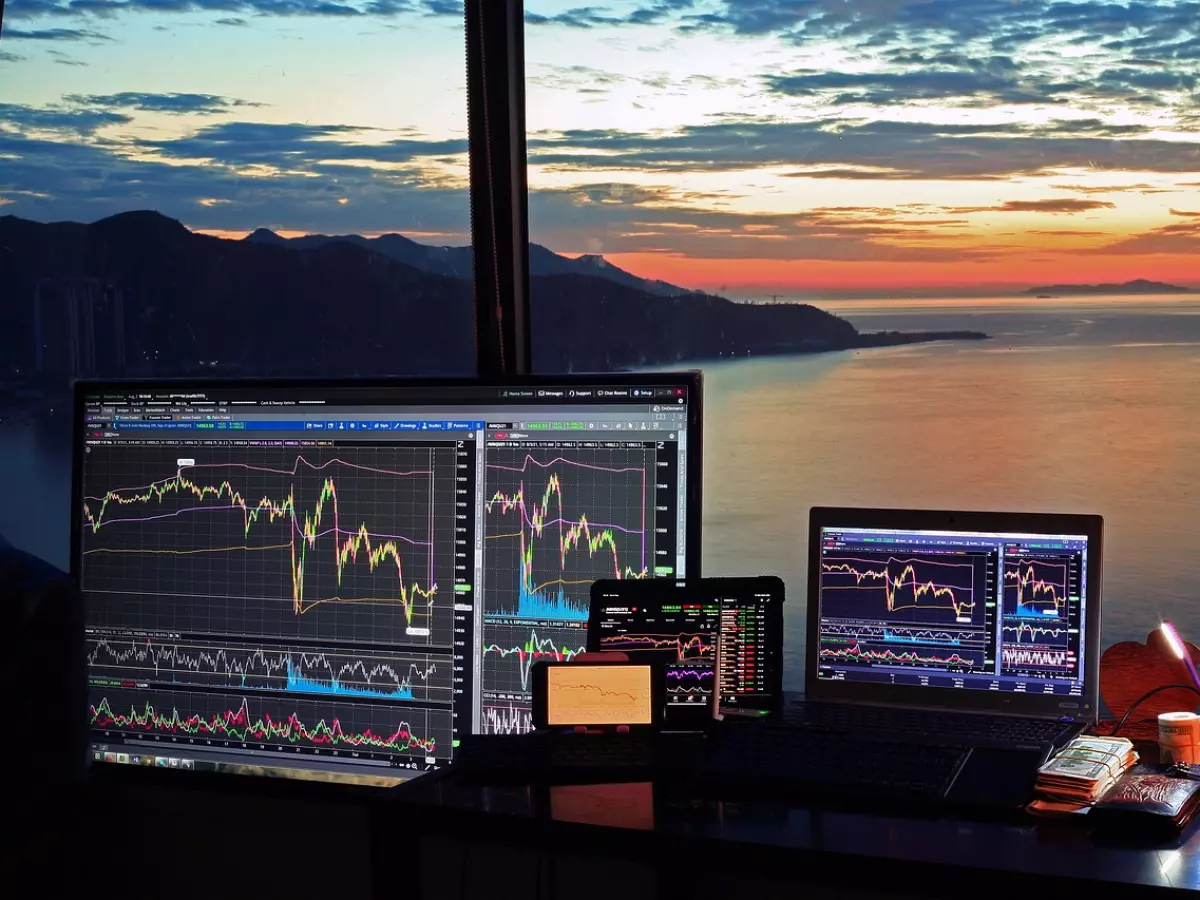 A desk with multiple devices displaying stock market charts. The scene is set in a room with a large window offering a panoramic view of a cityscape at sunset. The desk is also equipped with a keyboard, mouse, and other accessories, highlighting the digital nature of the subject matter.