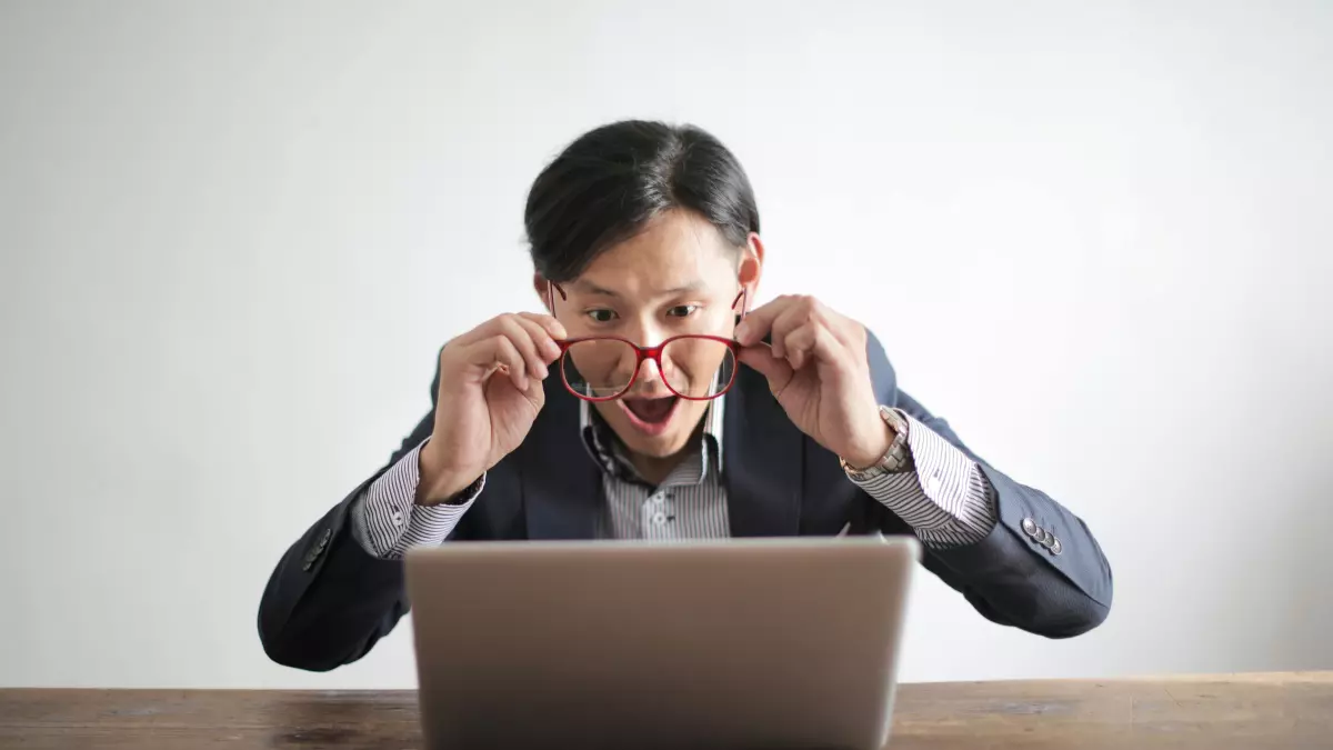 A man wearing glasses sits at a desk looking at his laptop with a surprised expression. He is holding his glasses with his hands.