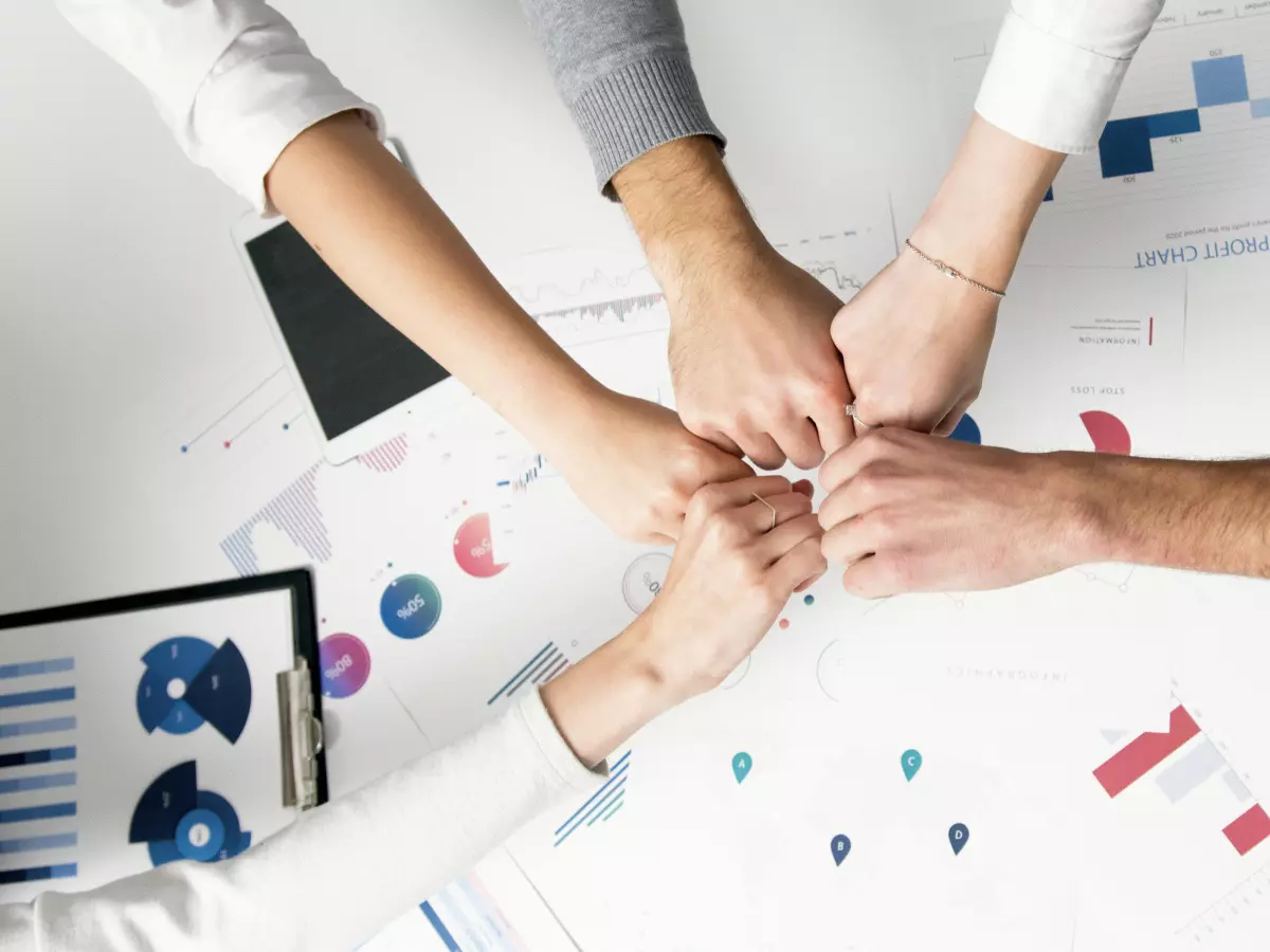 A group of five people are gathered around a table with their hands in the middle. The image is taken from above, and the table is covered in papers and charts. One of the people is holding a smartphone.