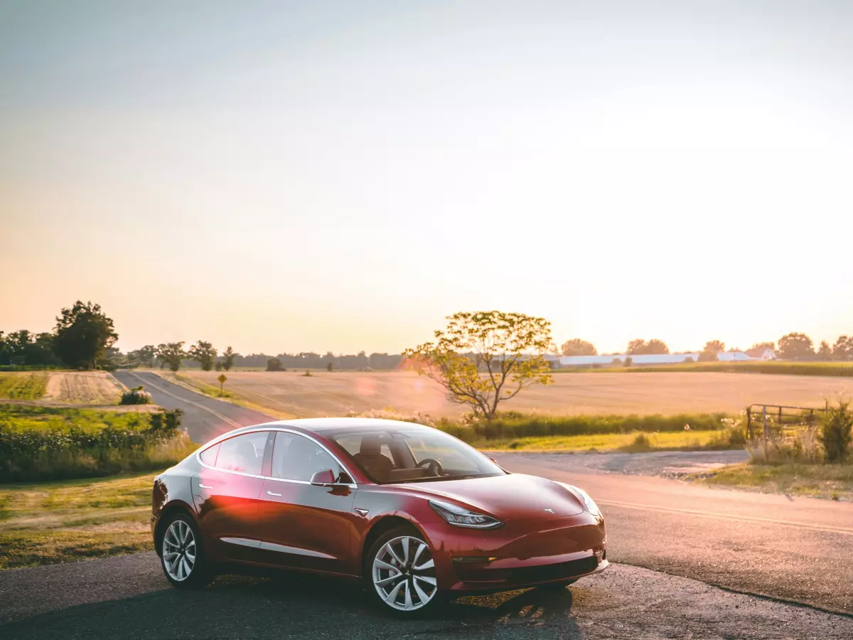 A red Tesla Model 3 driving on a country road at sunset, with a field and trees in the background.