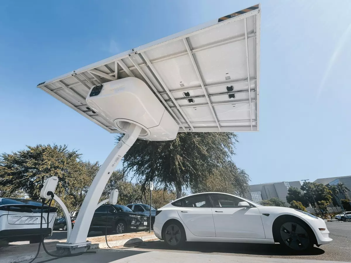 A solar-powered EV charging station with a white Tesla car plugged in. The station has a large solar panel on top and is located in a parking lot with other cars in the background.