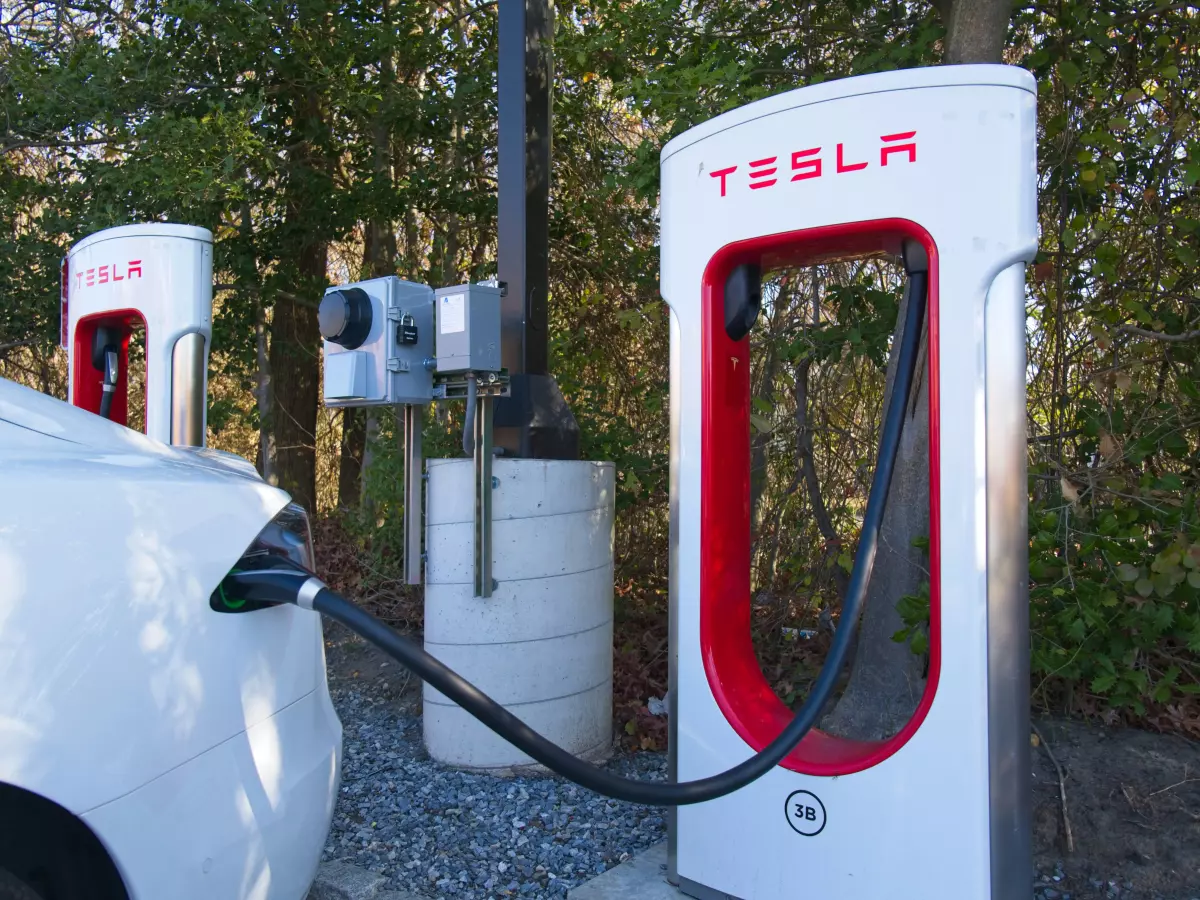 A Tesla Supercharger station with two charging stalls. One Tesla car is connected to the charging station and is being charged. Another Tesla car is parked behind it.