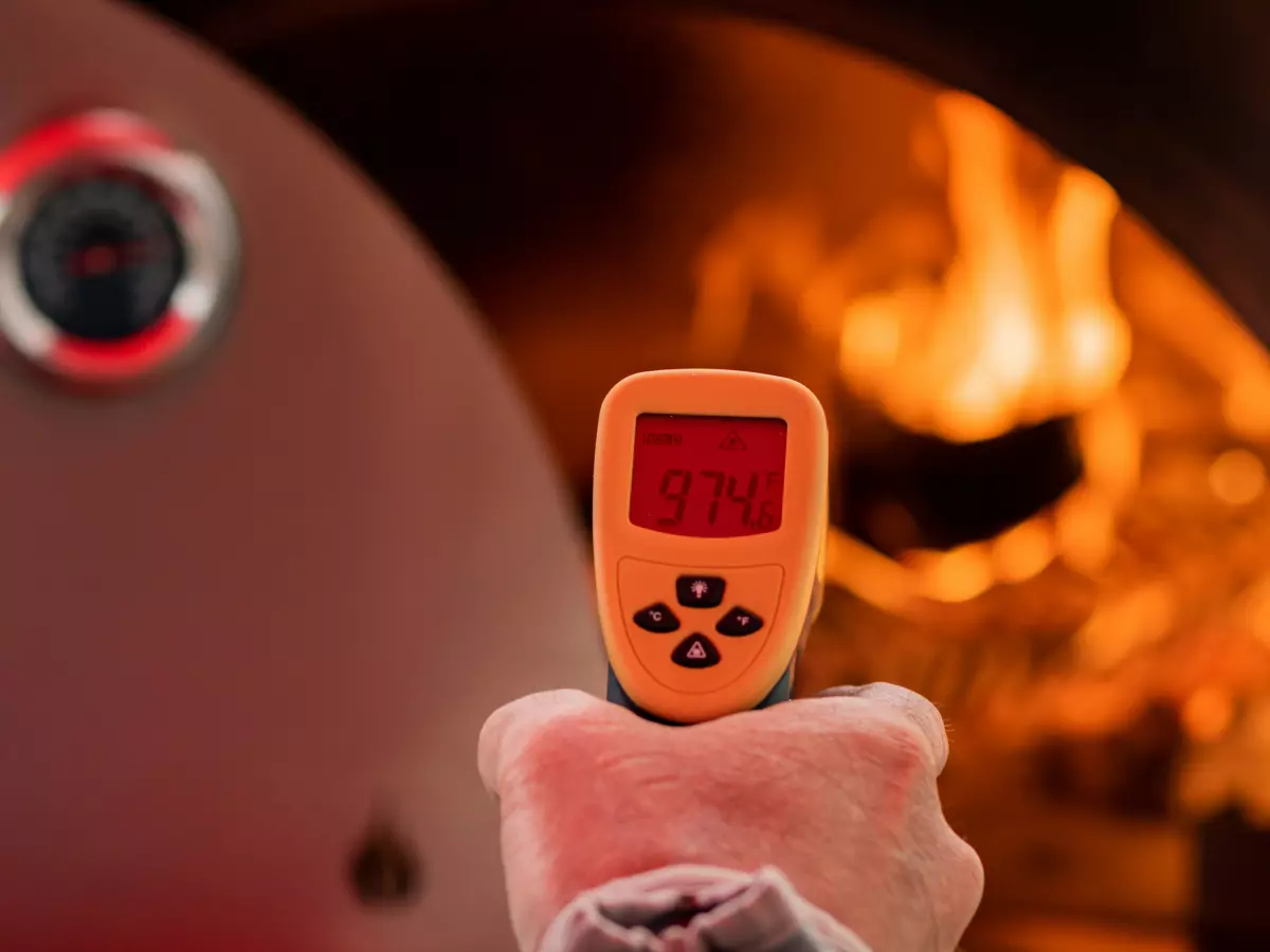 A hand holding a digital thermometer with a bright red display showing the temperature 97.4. The background is blurred with a fire in a pizza oven visible.