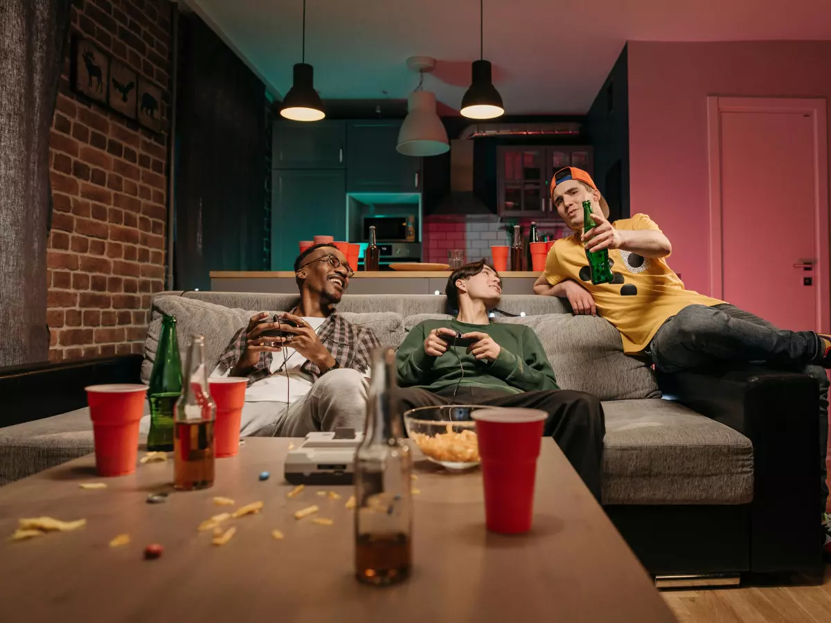 A group of friends are gathered in a living room playing video games, with one person on the couch using a controller and the others watching.