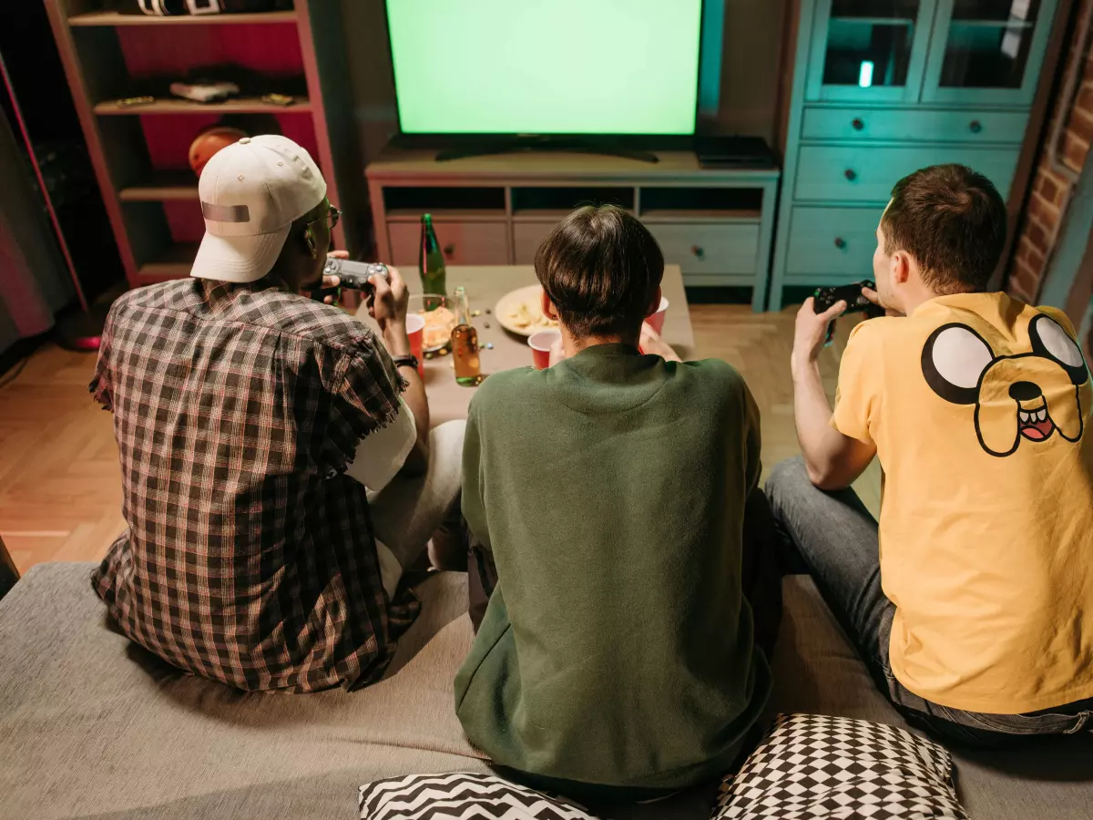 A group of three young men are playing a video game on a large screen TV in a living room. They are all sitting on the floor, and one is holding a controller. The image is taken from behind the men, and it captures their excited expressions as they play.