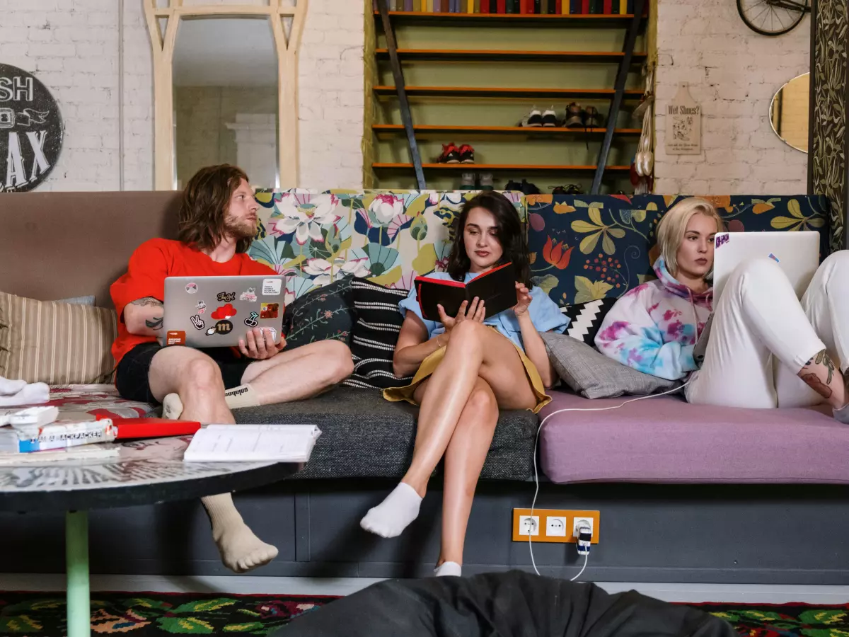 Three young people are sitting on a sofa, looking at their devices. One is using a laptop, another is reading a book, and the third is on their phone. The sofa has a floral pattern and the room is filled with bookshelves.