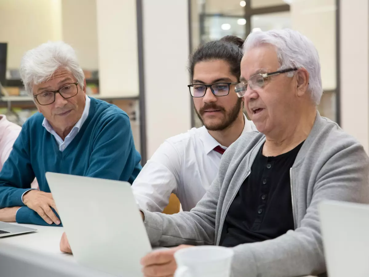 Three men are sitting at a table in a library, looking at a laptop computer.