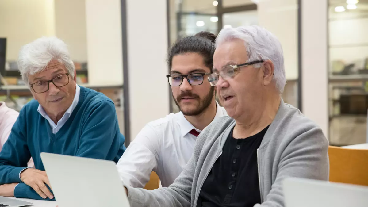 Three men are sitting at a table in a library, looking at a laptop computer.