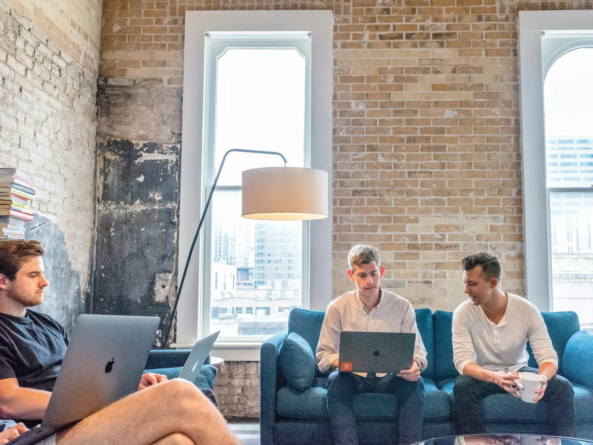 Three people sitting on a couch in a loft apartment, discussing something on their laptops.