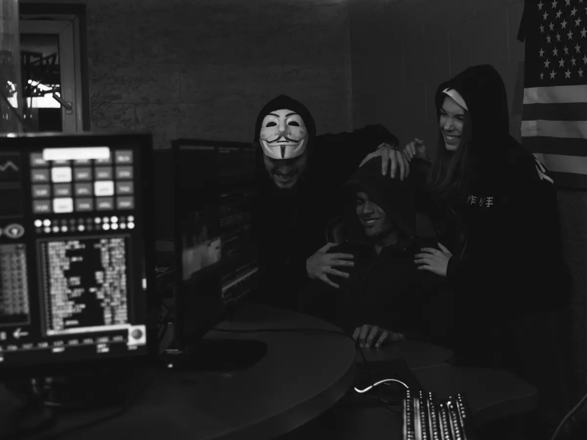 Two masked hackers in a dimly lit room, sitting in front of a computer with a US flag in the background.