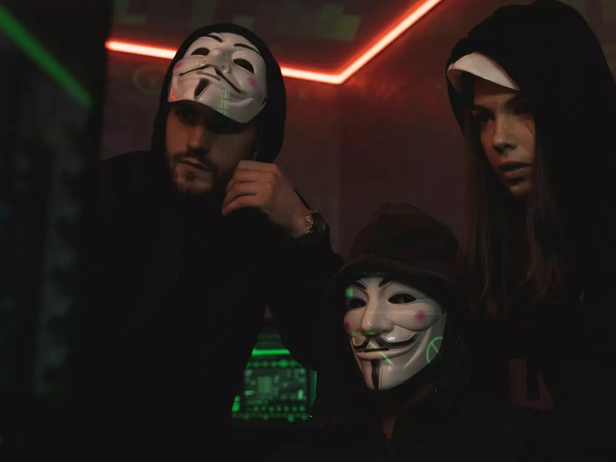 Three people wearing black hoodies and masks are looking at a computer screen, the image is shot from a low angle.