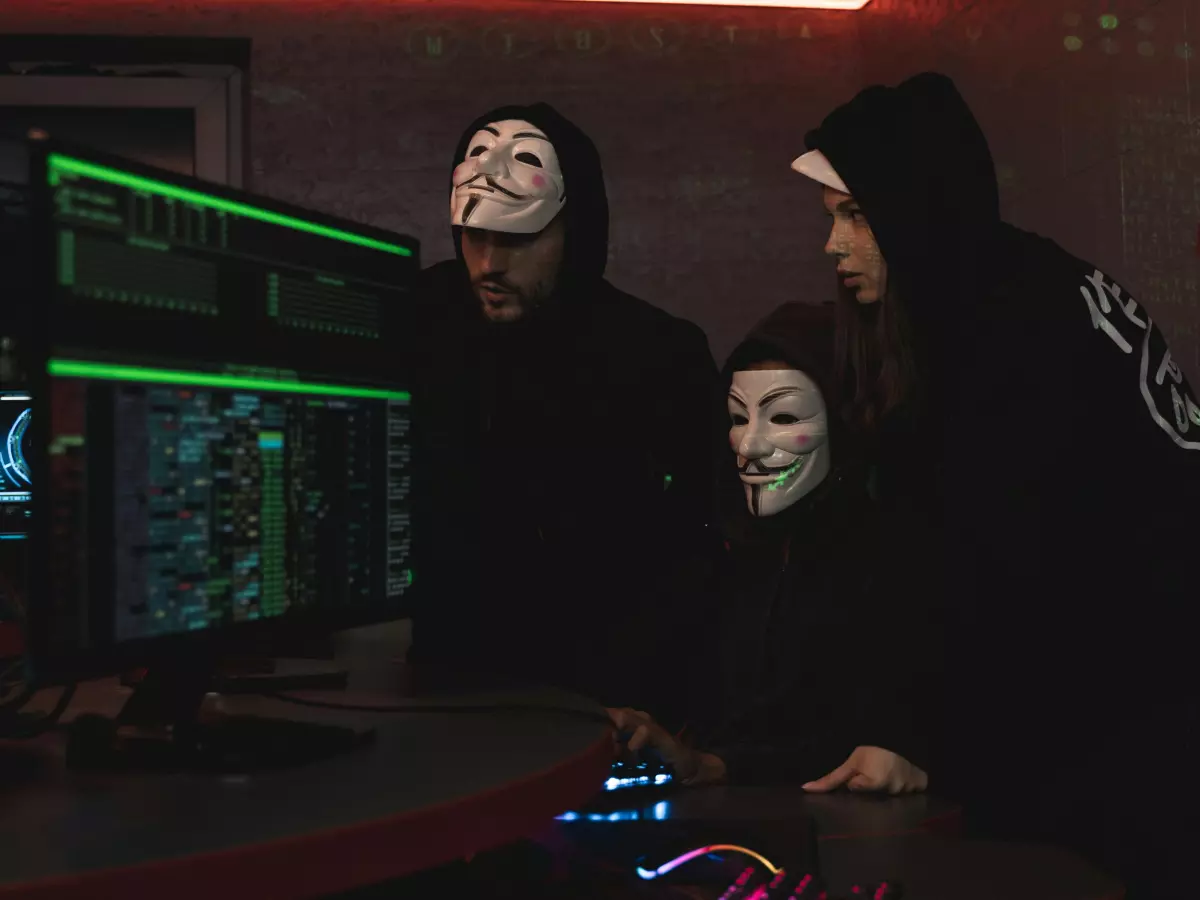 Three masked individuals are hunched over a computer, engaged in a seemingly illicit activity.  The green screens behind them depict code, further emphasizing their sinister intent. The room is dark and dimly lit, contributing to the overall sense of secrecy and danger.