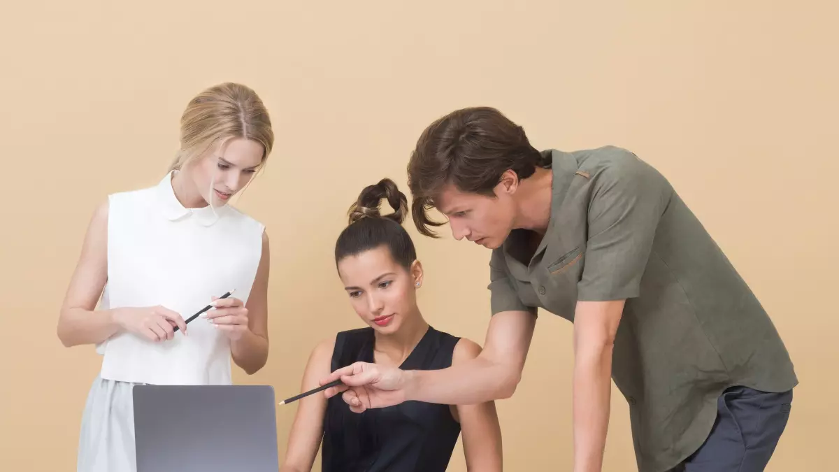 Three people working on a laptop together in an office setting. The man in the back is leaning in and pointing at the laptop screen. The two women are looking at the screen and listening to him. The atmosphere is professional and collaborative.