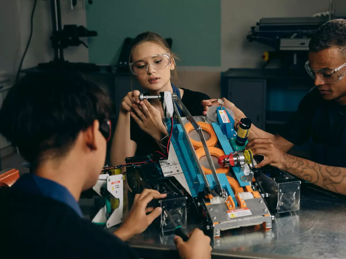 A group of people working on a robot, the focus is on a woman's face, she looks up and has a thoughtful expression. There is a robot arm in the middle of the table.