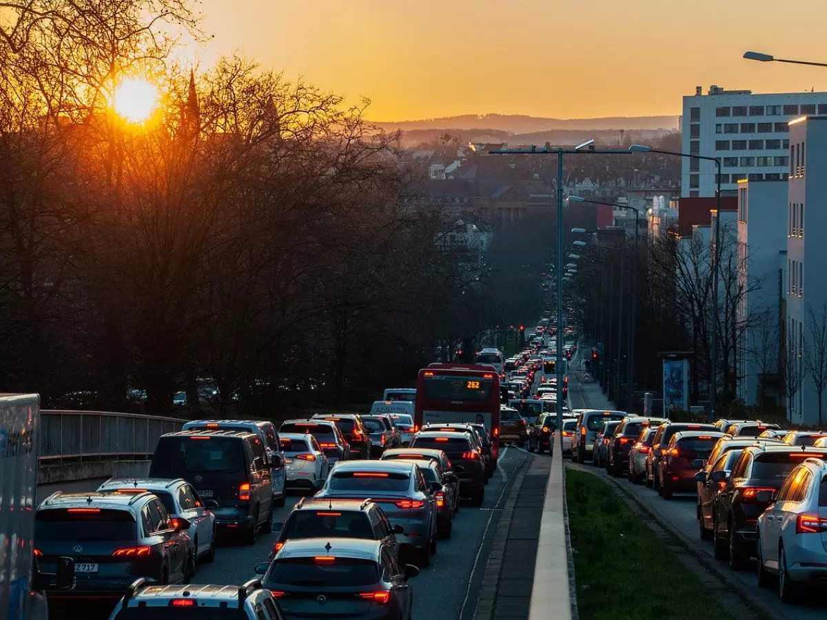 A long line of cars stuck in a traffic jam on a city street during sunset. The sun is setting behind the buildings in the distance.