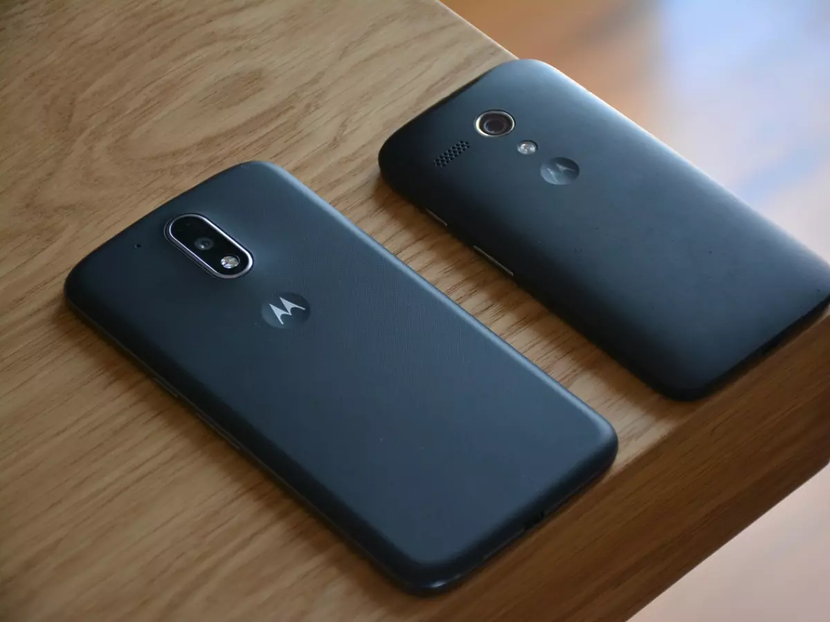 Two Motorola smartphones, one black and one dark gray, are shown on a wooden surface. The smartphones are placed next to each other, with the black one on the right and the gray one on the left. The image is taken from a top-down perspective, showing the back of the phones.