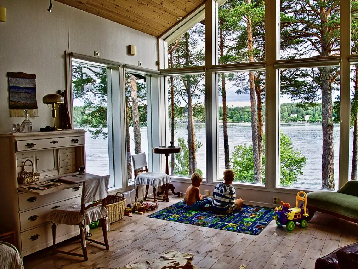 A living room with large windows looking out onto a lake. Two children are sitting on the floor looking out the window. There is a white dresser with a lamp on top and a green chaise lounge in the room.