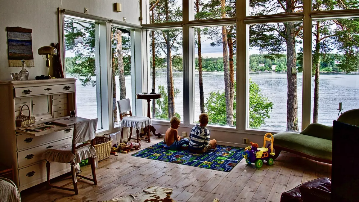 A living room with large windows looking out onto a lake. Two children are sitting on the floor looking out the window. There is a white dresser with a lamp on top and a green chaise lounge in the room.