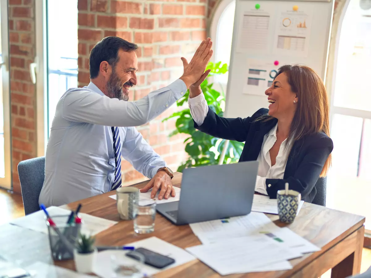 Two professionals in a business setting, a man and a woman, give each other a high-five while sitting at a table with a laptop.