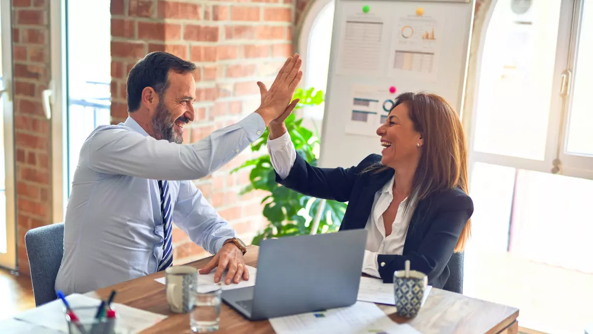 Two professionals in a business setting, a man and a woman, give each other a high-five while sitting at a table with a laptop.