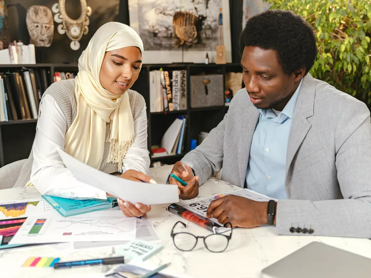 Two people, a woman in a headscarf and a man, are sitting at a table and looking at papers, both of them appear engaged in a project.