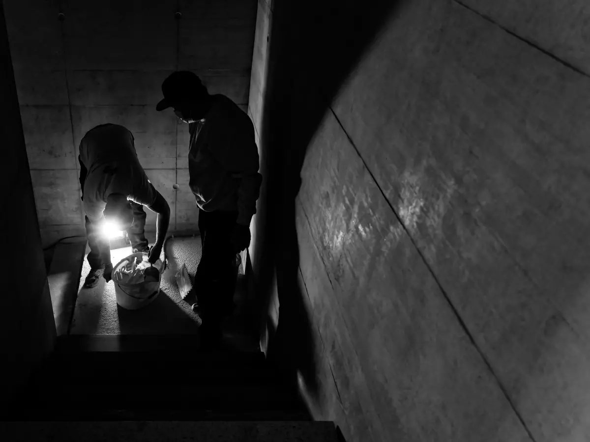 Two people working on a construction site, one holding a bucket, the other holding a light.