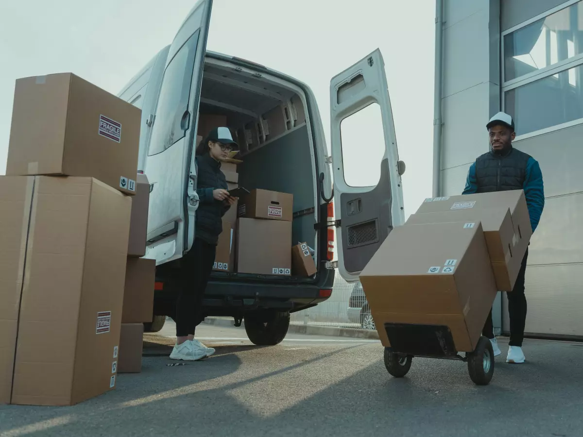 A delivery van being loaded with boxes. There are two people, one inside the van and one loading boxes from a dolly. The background is a loading dock.