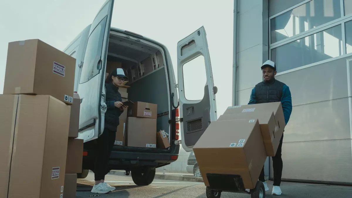 A delivery van being loaded with boxes. There are two people, one inside the van and one loading boxes from a dolly. The background is a loading dock.