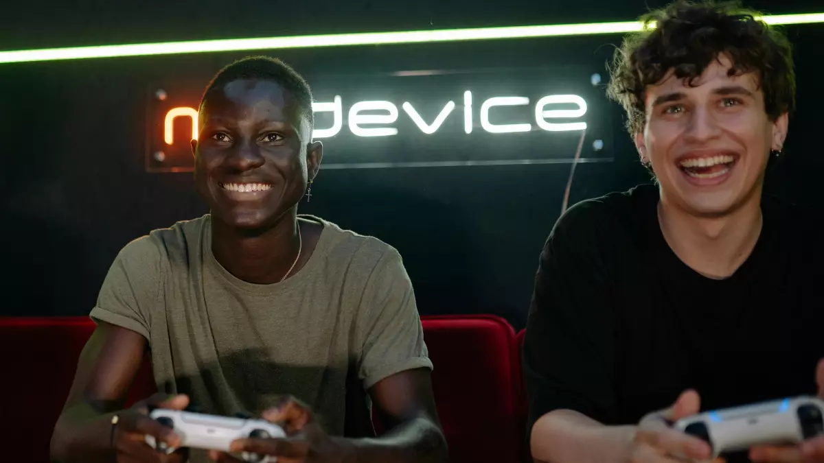 Two men are sitting on a couch and playing video games with controllers. They are both smiling and having a good time. The background is a bit dark, but the men are well-lit and the controllers are very visible. The image is inviting and engaging.