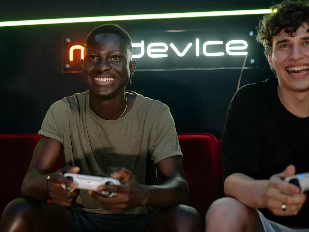 Two men are sitting on a couch and playing video games with controllers. They are both smiling and having a good time. The background is a bit dark, but the men are well-lit and the controllers are very visible. The image is inviting and engaging.