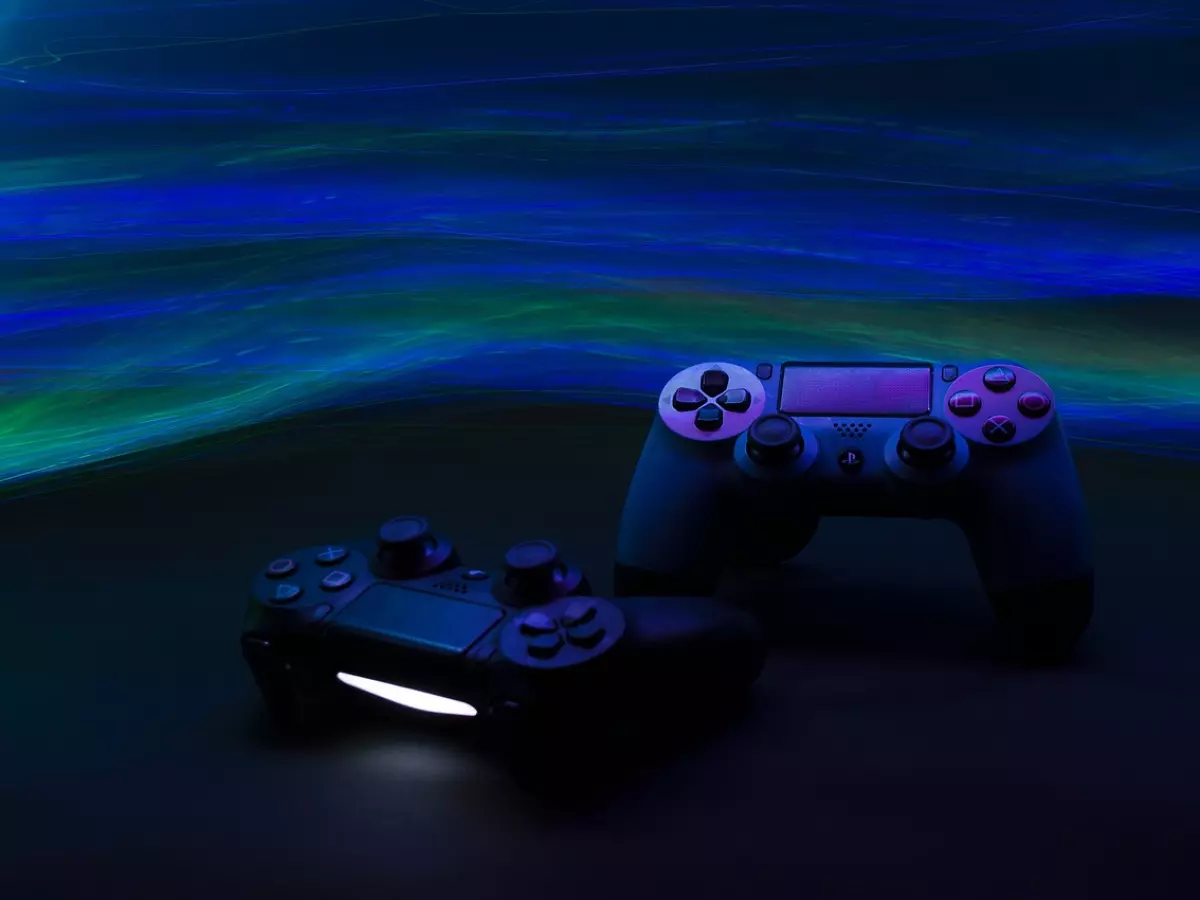 Two PS5 controllers, one black and one blue, are placed on a table with a blue and green light behind them.