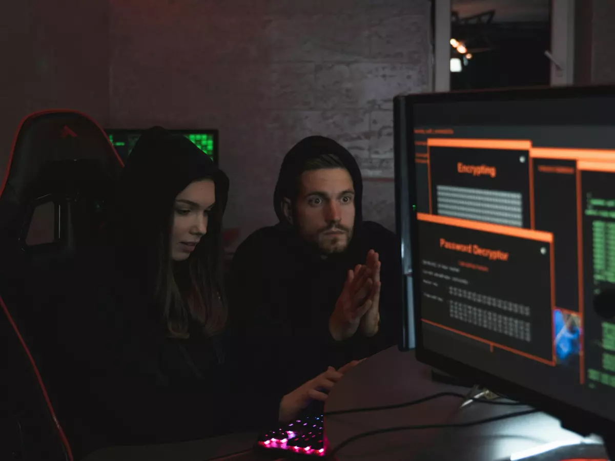 Two hackers in hoodies are working on a computer in a dimly lit room. The image is full of code, with a mysterious aura surrounding them. 