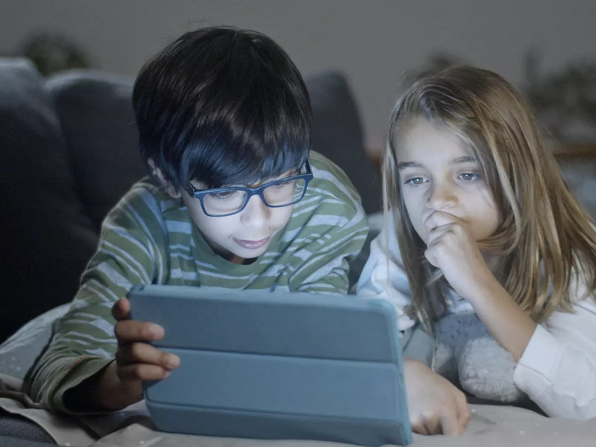 Two young children, a boy and a girl, are lying on a couch and using a tablet computer. They are both looking at the screen intently. 